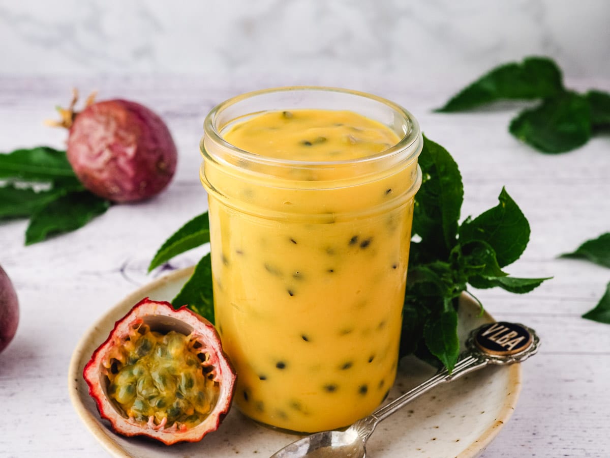 Mason jar of curd without lid on a plate with spoon and fresh passion fruit and leaves on the side.