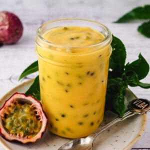 Close up mason jar of curd without lid on a plate with spoon and fresh passion fruit and leaves on the side.