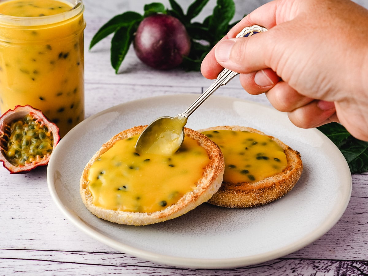 Spooning curd onto English muffins on a plate, with jar of curd and fresh passion fruit in background.