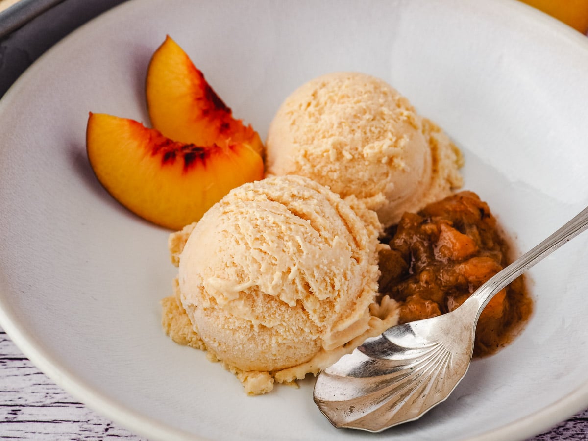 Two scoops of ice cream in a bowl with peach compote, fresh peach slices and a spoon.