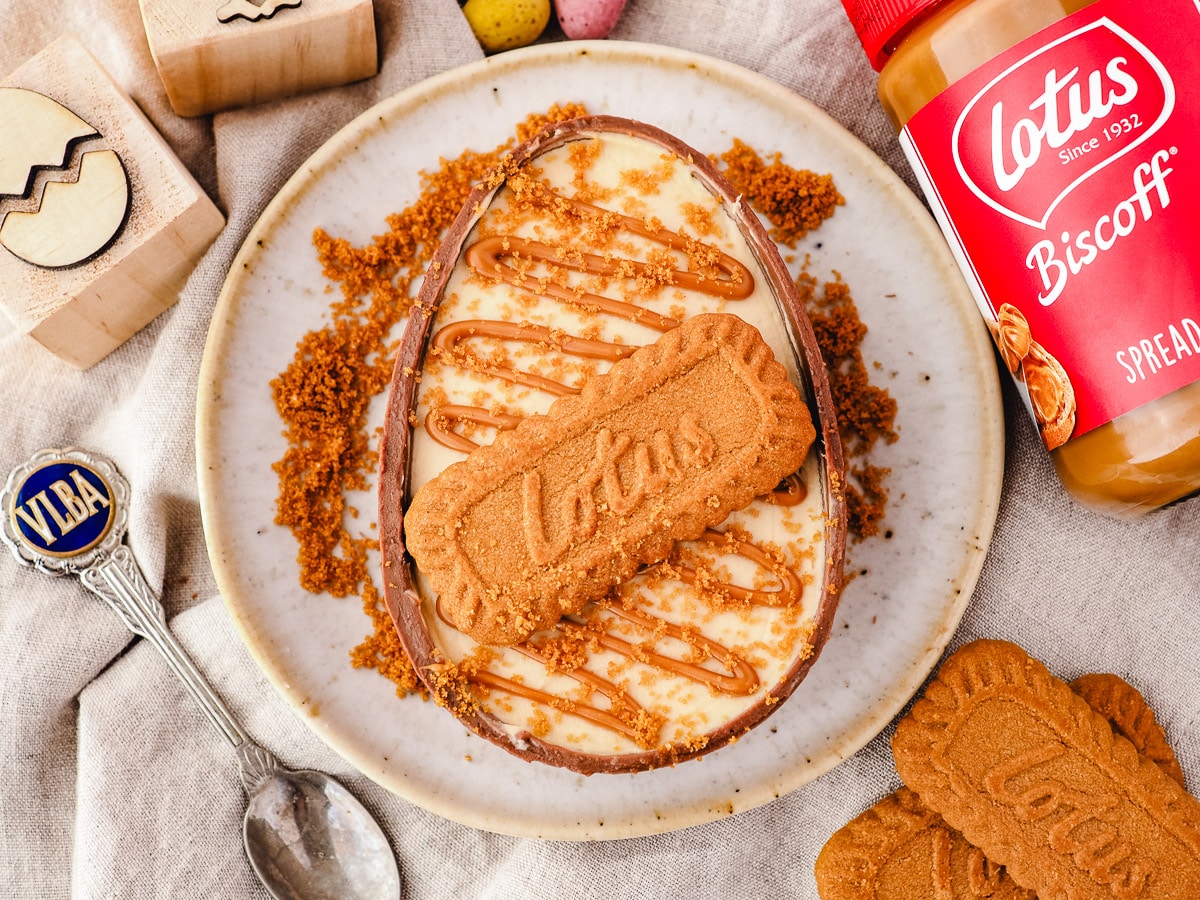 Large Biscoff cheesecake in Easter egg on a plate, surrounded by crumbs, with spoon and jar of Biscoff spread on the side.