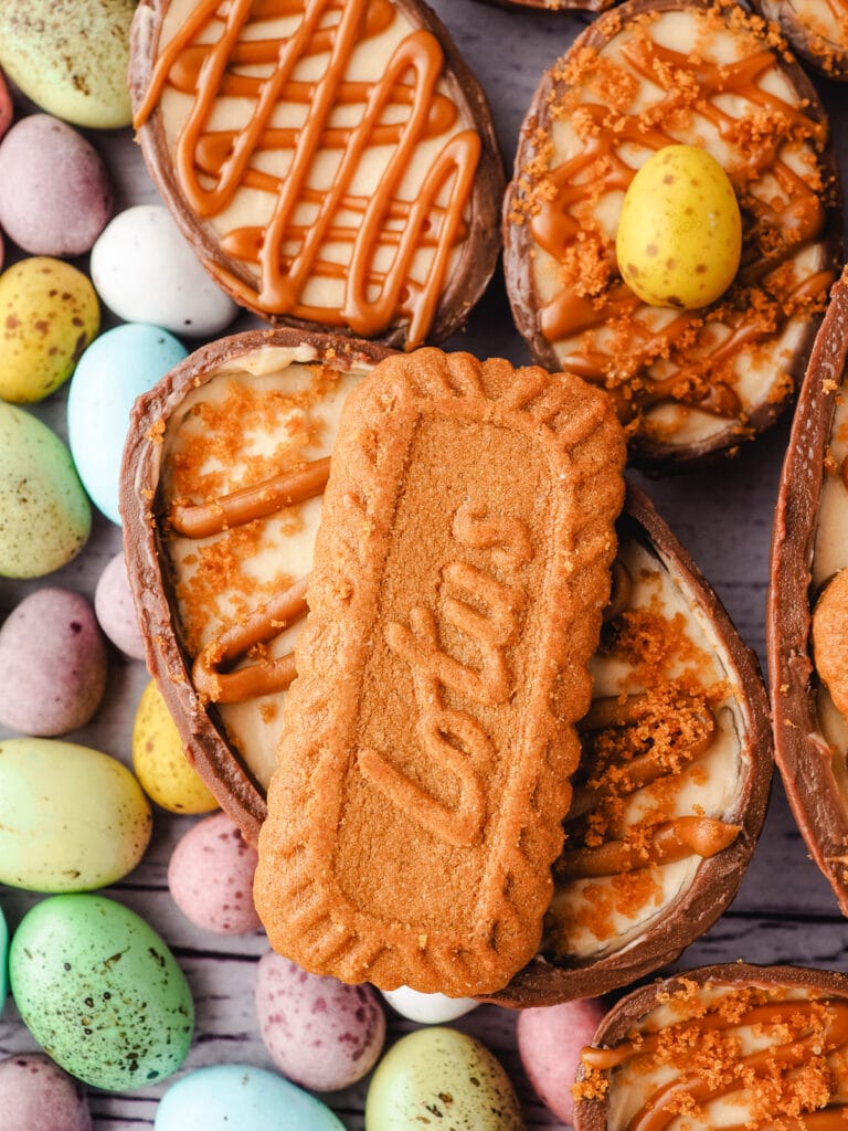 Biscoff Easter Egg Cheesecake 12 Cheesecake drizzled with spread, whole biscuit and crumbs, with colorful mini eggs on the side.