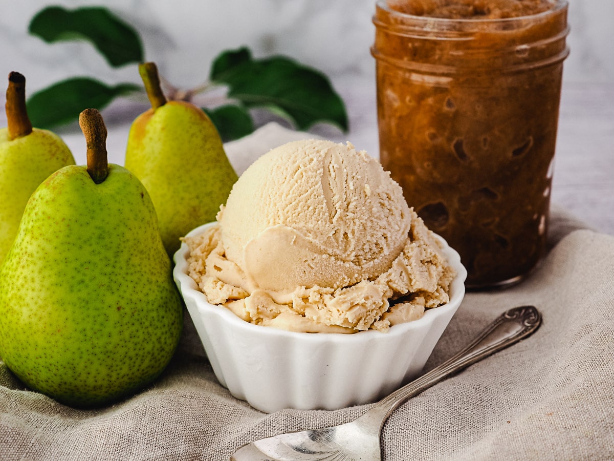 Scoop of pear ice cream with spoon, fresh pears and jar of pear compote on the side.