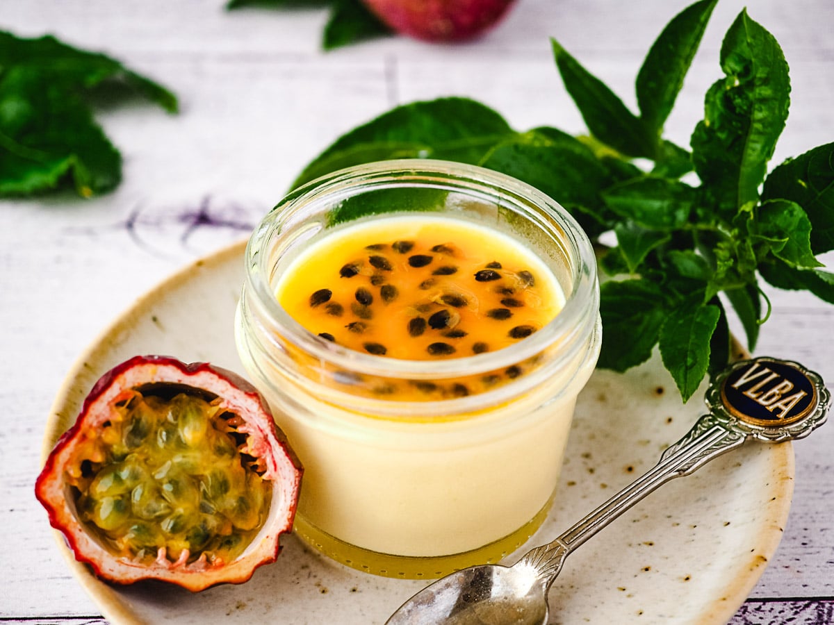 Small mason jar of panna cotta with passion fruit coulis, on a plate with a spoon and fresh passion fruit.