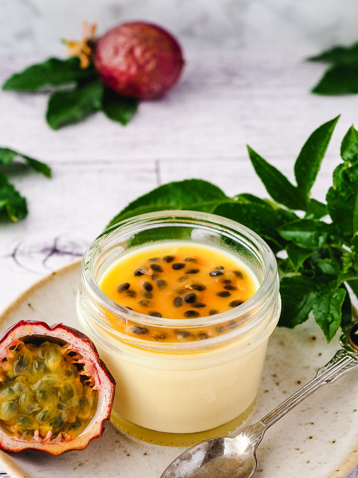 Small mason jar of panna cotta with passion fruit coulis, on a plate with a spoon and fresh passion fruit.