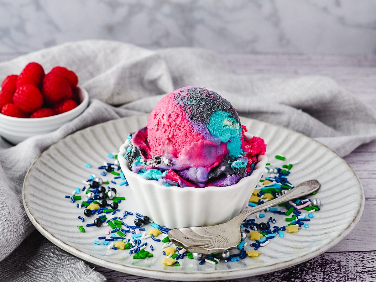 Scoop of ice cream with sprinkles, on a plate with spoon and sprinkles, and raspberries in the background.