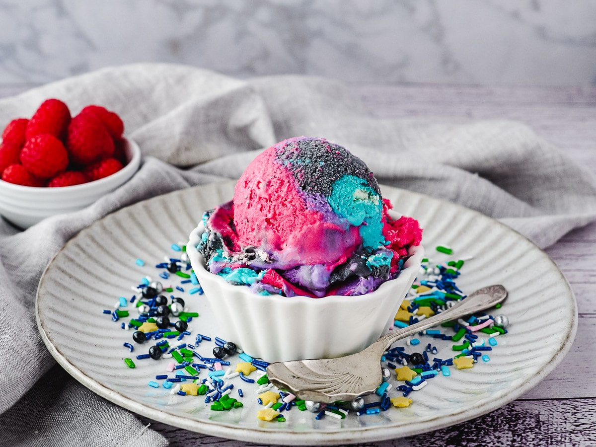 Galaxy Ice Cream 7 Scoop of ice cream with sprinkles, on a plate with spoon and sprinkles, and raspberries in the background.