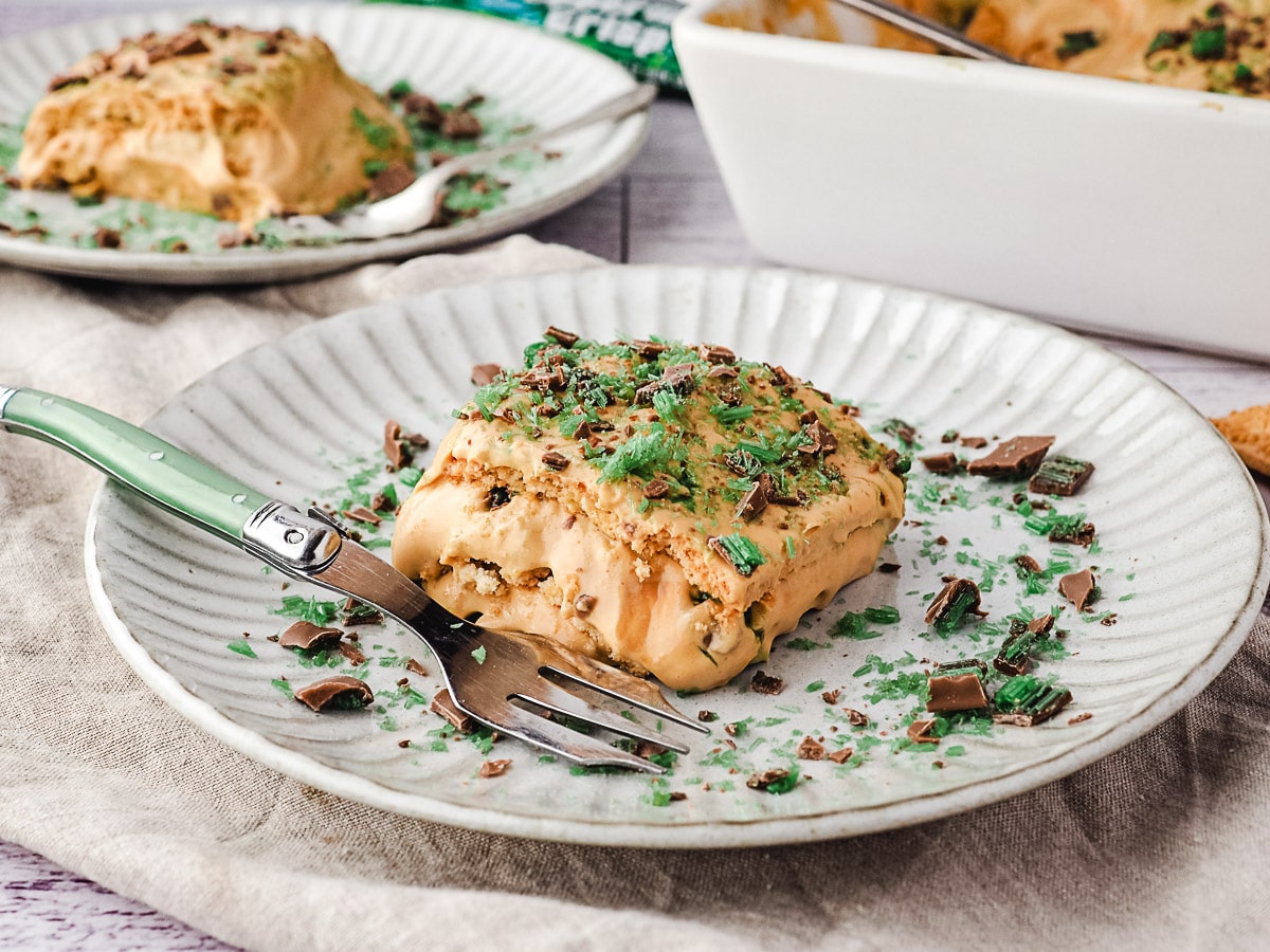 Slice of tart on a plate with a fork, with an extra slice of tart, rest of tart in baking tray and peppermint crisp bars in the background.