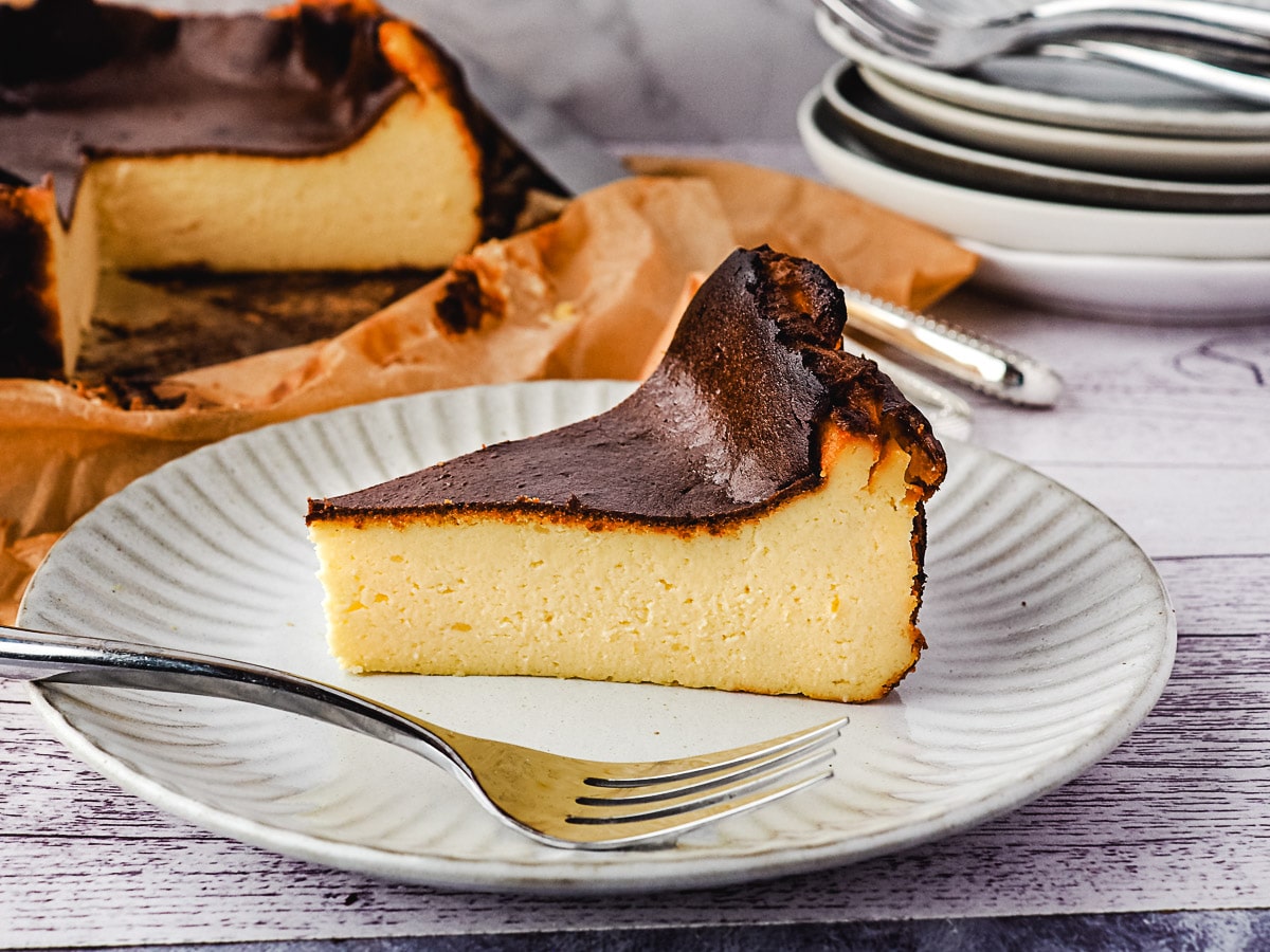 Slice of cheesecake on a plate with fork and rest of cheesecake and stack of plates and forks in the background.