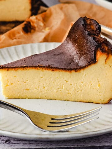 Slice of cheesecake on a plate with fork and rest of cheesecake and stack of plates and forks in the background.