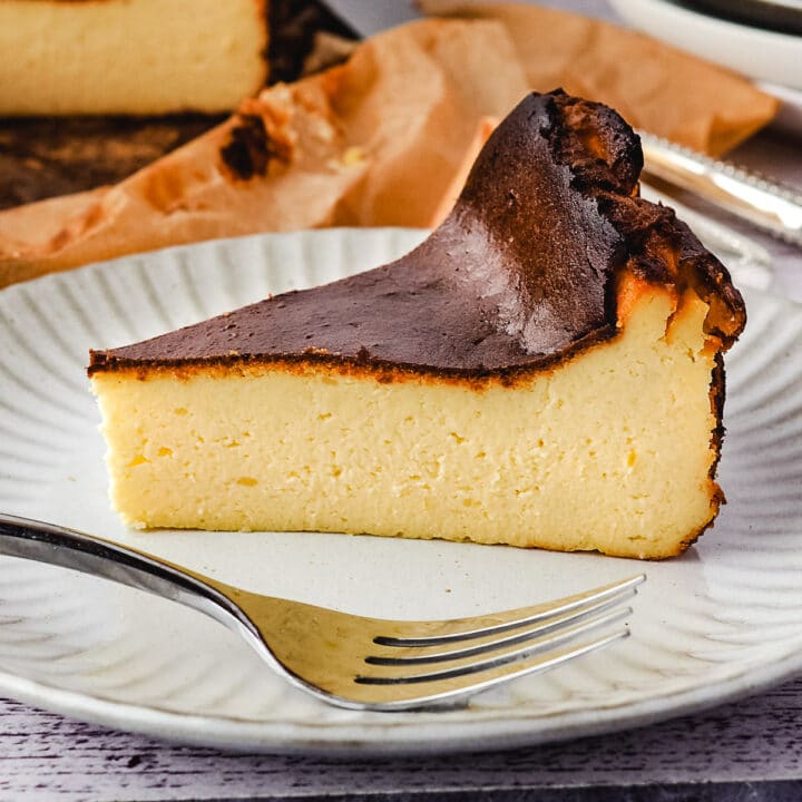 Slice of cheesecake on a plate with fork and rest of cheesecake and stack of plates and forks in the background.