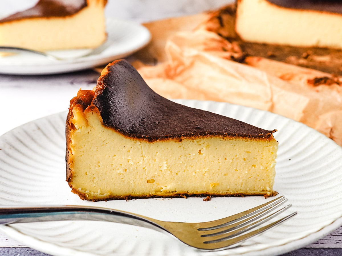 Slice of cheesecake on a plate with fork, with another slice of cake and rest of cheesecake in the background.