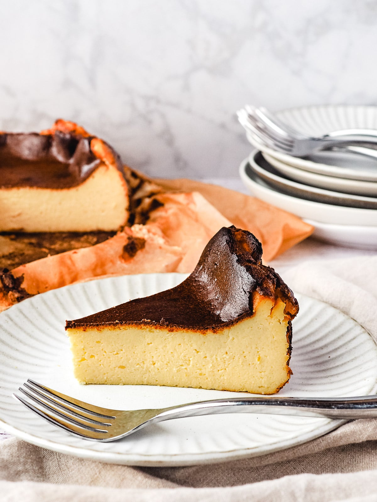 Slice of cheesecake on a plate with fork and rest of cheesecake and stack of plates and forks in the background.