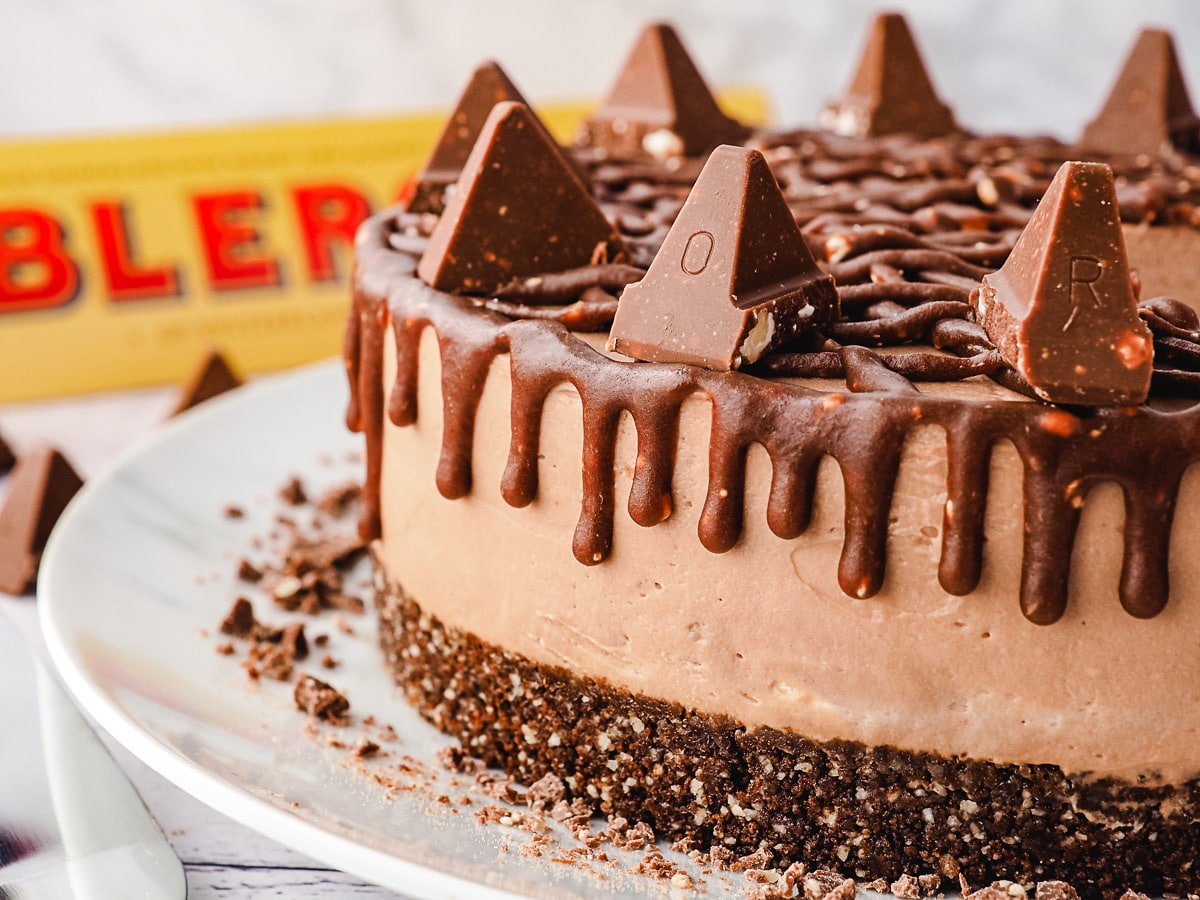 Close-up side view of whole cheesecake showing drip, with Toblerone in background.