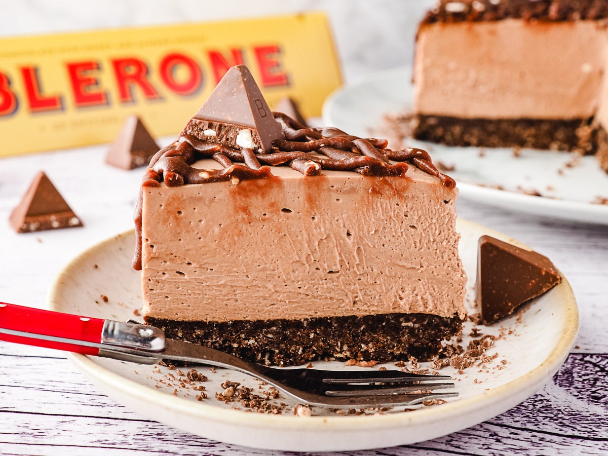 Slice of Toblerone cheesecake on a plate, with rest of cheesecake and more Toblerone in background.