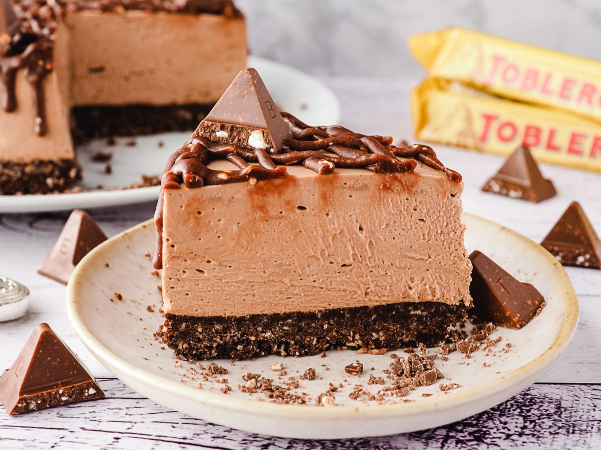 Slice of Toblerone cheesecake on a plate, with rest of cheesecake and more Toblerone in background.