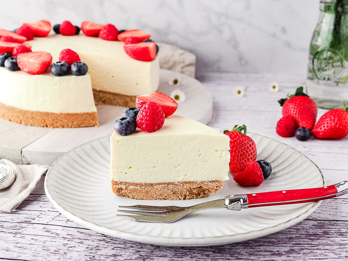 Slice of cheesecake on a plate with fresh berries and rest of cake and a small vase of flowers in the background.