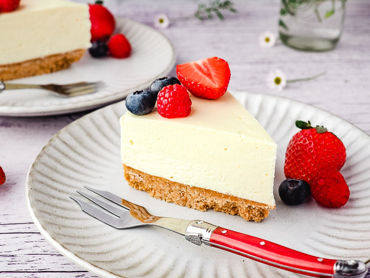 Slice of cheesecake on a plate with fresh berries, with another slice of cheesecake and small vase of flowers in the background.