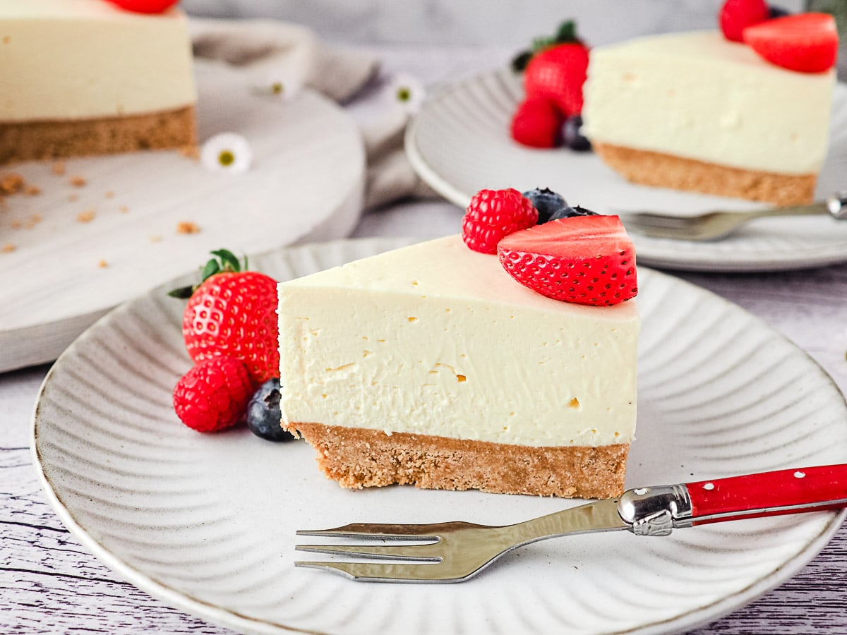 Slice of cheesecake on a plate with fresh berries and rest of cake and another slice of cheesecake in the background.
