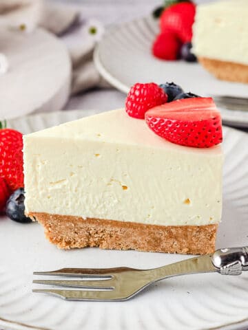 Slice of cheesecake on a plate with fresh berries and rest of cake and another slice of cheesecake in the background.