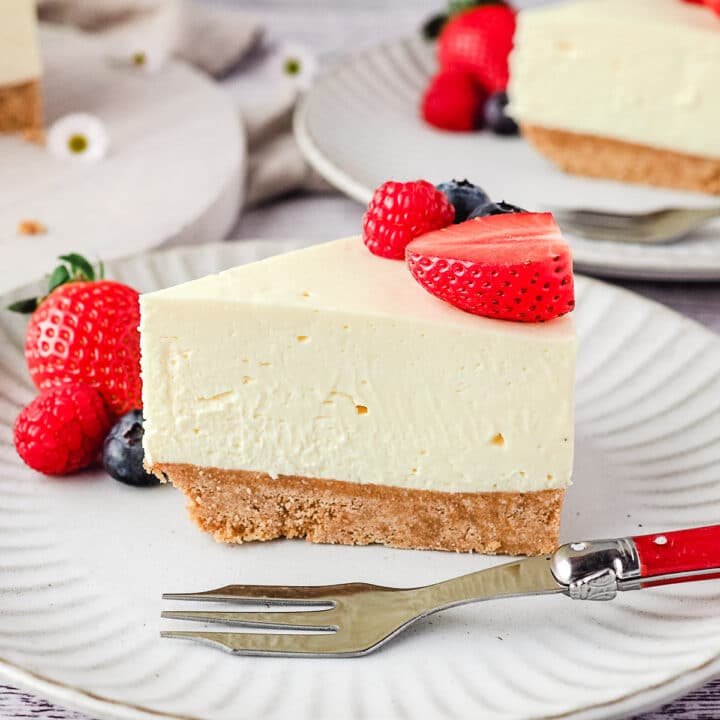 Slice of cheesecake on a plate with fresh berries and rest of cake and another slice of cheesecake in the background.