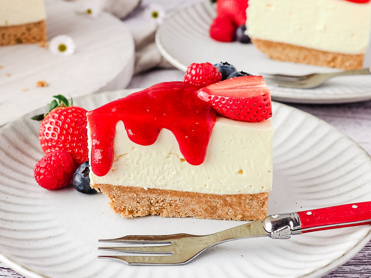 Slice of cheesecake on a plate with fresh berries and drizzle of strawberry sauce, and rest of cake and another slice of cheesecake in the background.