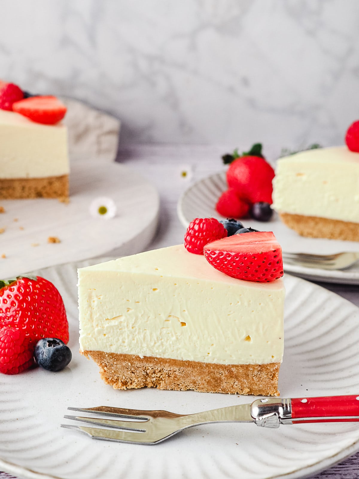 Slice of cheesecake on a plate with fresh berries and rest of cake and another slice of cheesecake in the background.