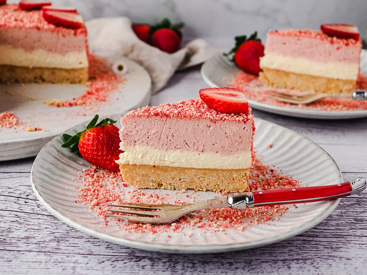 Strawberry Crunch Cheesecake 2 Slice of cheesecake on a plate with a fork, and another slice of cake and rest of cake in background.