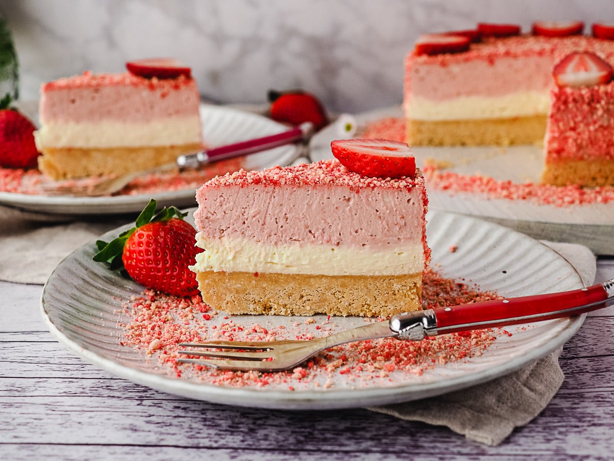 Slice of cheesecake on a plate with a fork, and another slice of cake and rest of cake in background.