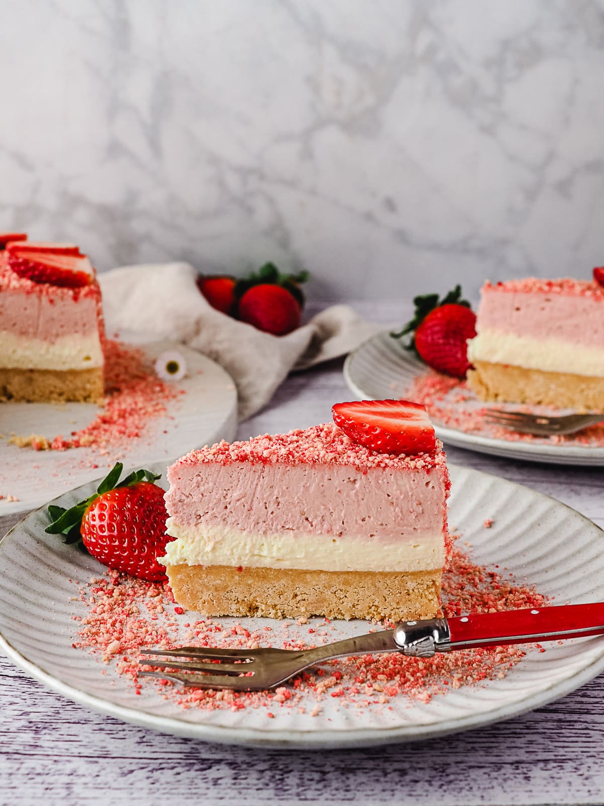 Slice of cheesecake on a plate with a fork, and another slice of cake and rest of cake in background.