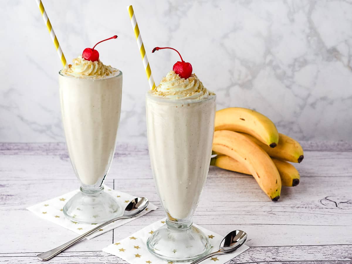 Two milkshakes in milkshake glasses, with straws, whipped cream, banana chip crumbs and a cherry on top, with fresh bananas in the background.