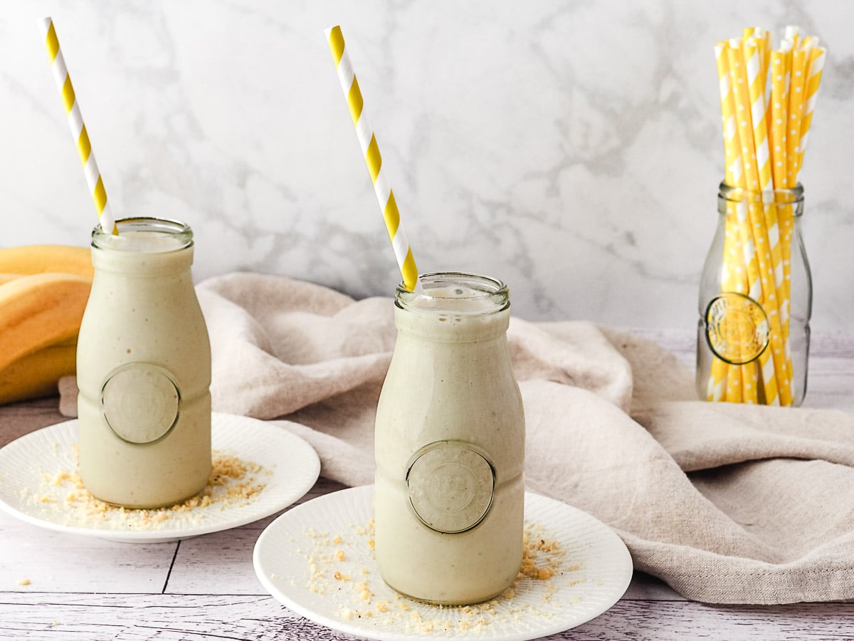 Two milkshakes with a straw in jars, with fresh bananas and jar of straws in background.