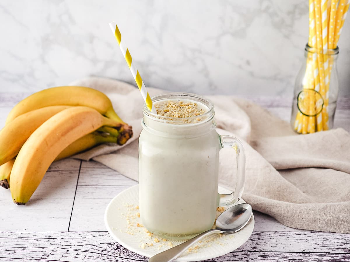 Milkshake in a mason jar with a straw and banana chip crumbs on top, with fresh bananas and jar of straws in background.