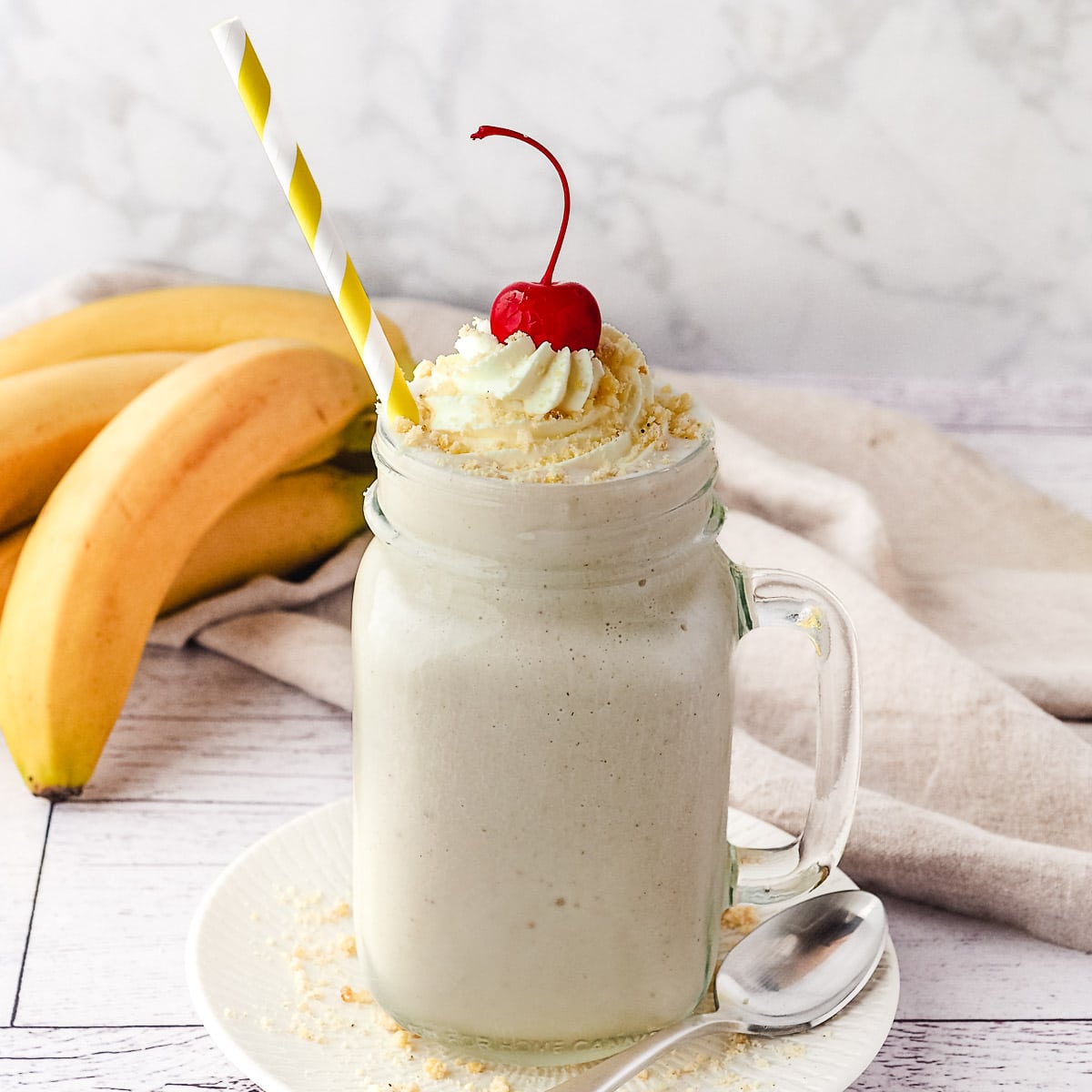 Milkshake in a mason jar with a straw, whipped cream, banana chip crumbs and a cherry on top.