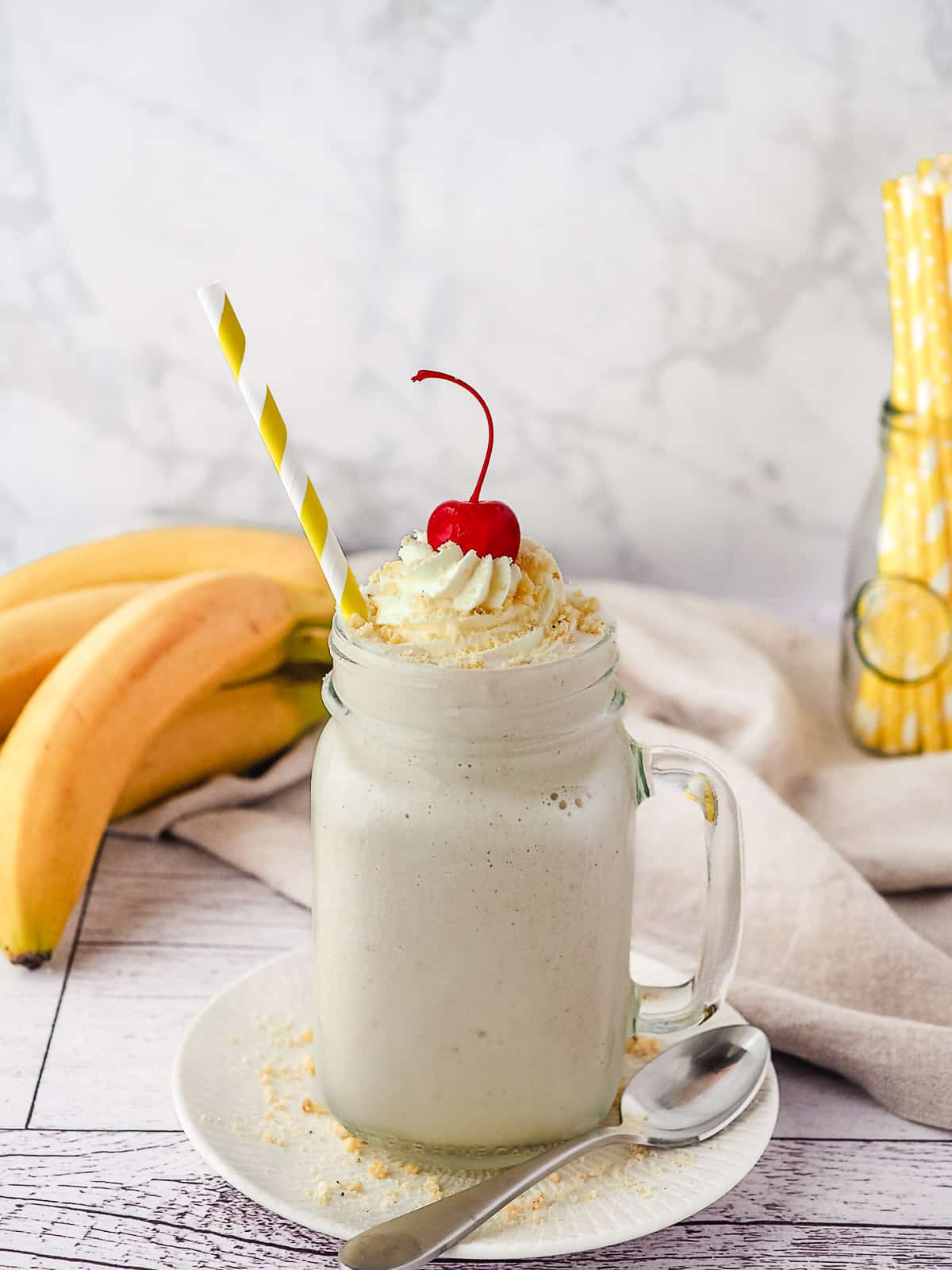 Milkshake with a straw, whipped cream, banana chip crumbs and a cherry on top, with fresh bananas and jar of straws in background.