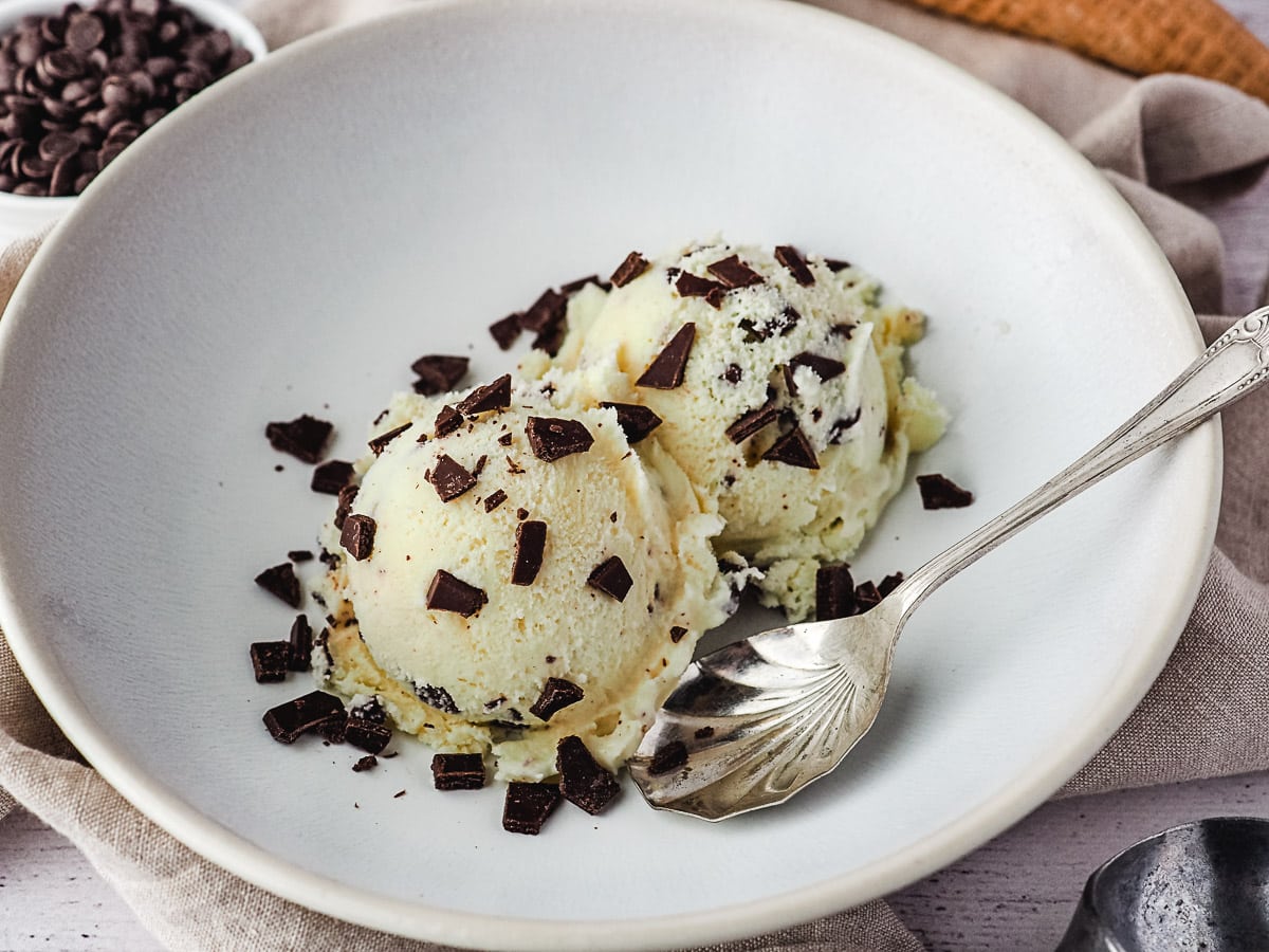 Two scoops of ice cream in a white bowl sprinkled with extra choc chips, a vintage spoon on the side, ice cream cones and bowl of choc chips in the background.