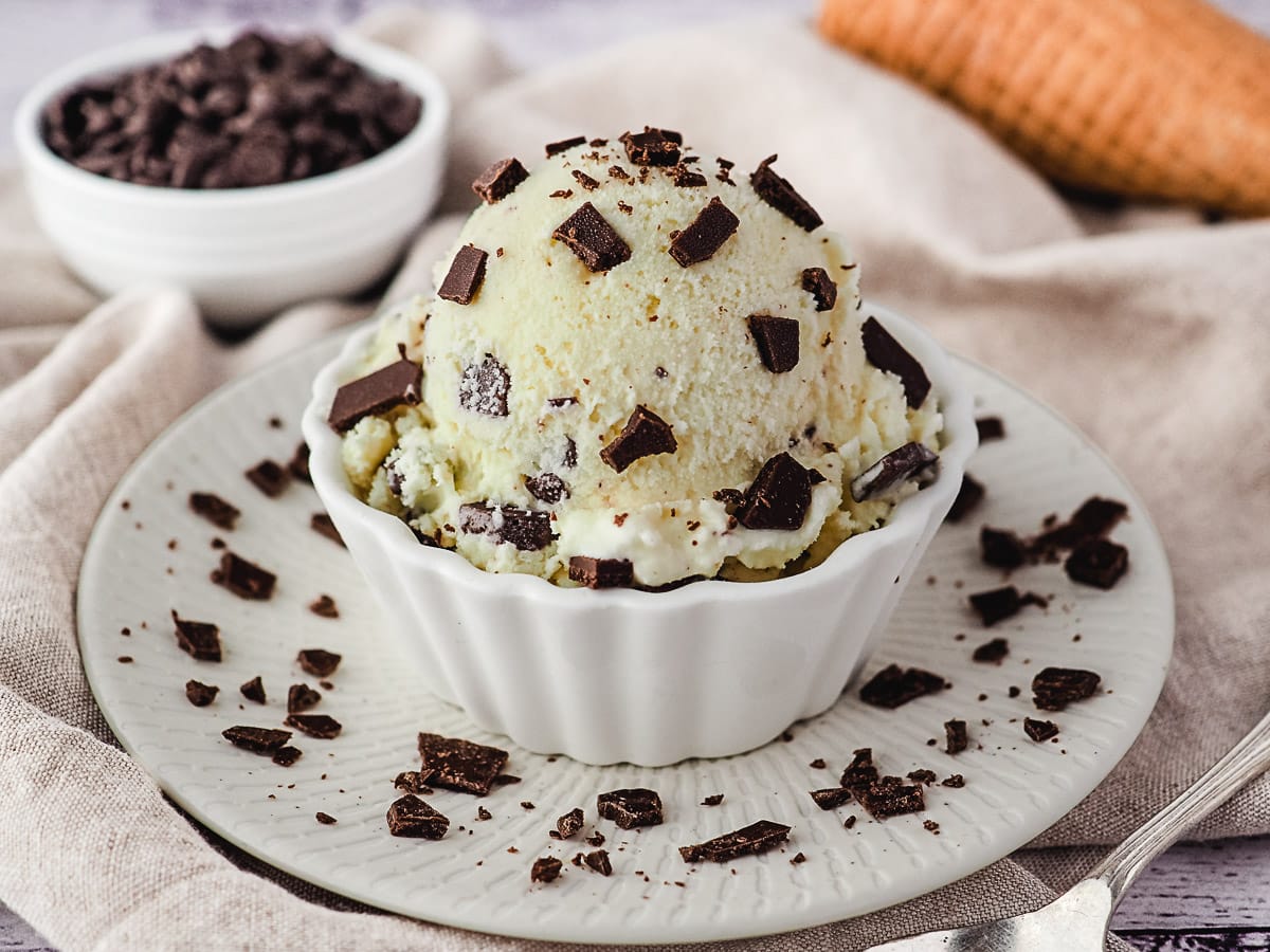 Scoop of ice cream in a bowl sprinkled with extra chocolate chips, and ice cream cones and bowl of choc chips in the background.