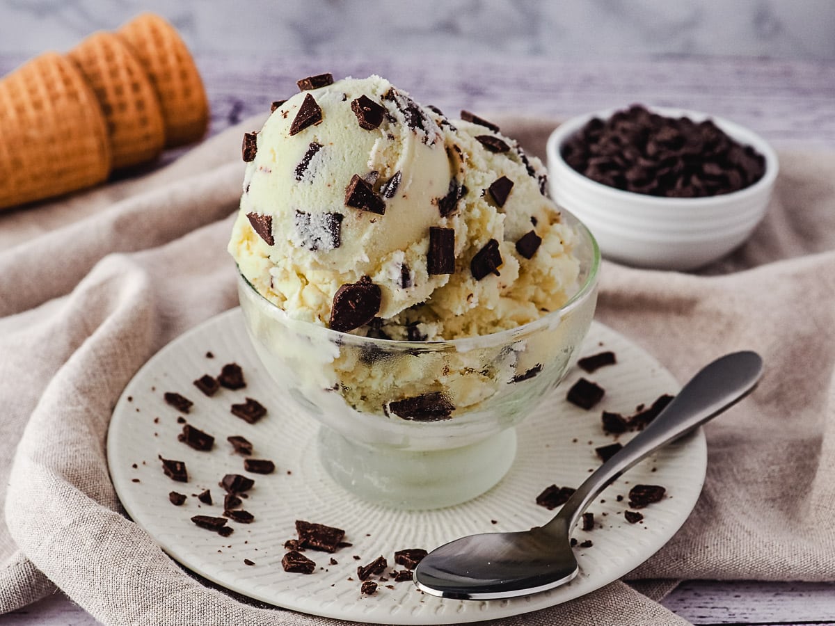 Two scoops of ice cream in a clean bowl sprinkled with extra chocolate chips, and ice cream cones and bowl of choc chips in the background.