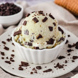 Close up scoop of ice cream in a bowl sprinkled with extra chocolate chips, and ice cream cones and bowl of choc chips in the background.