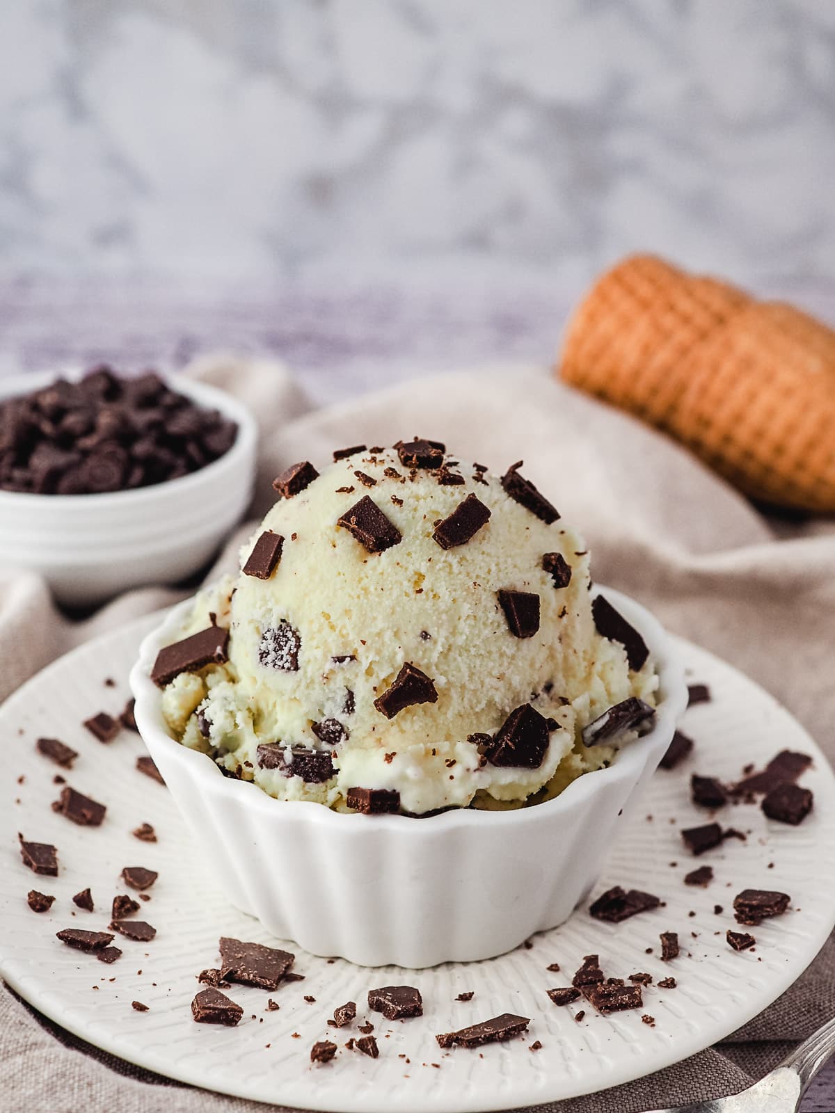 Scoop of ice cream in a bowl sprinkled with extra chocolate chips, and ice cream cones and bowl of choc chips in the background.