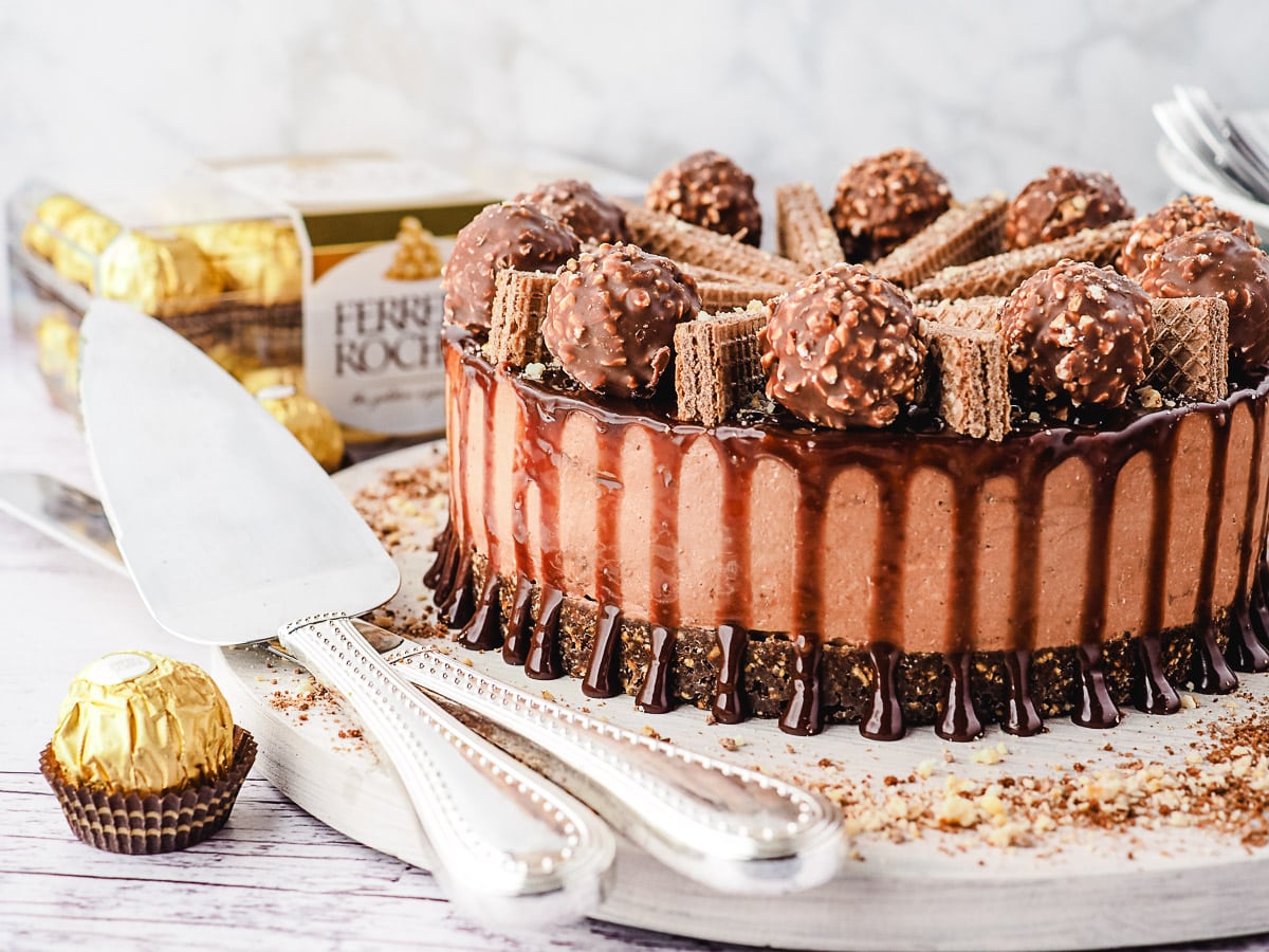 Cheesecake on a board with serving ware and Ferrero Rocher in background.