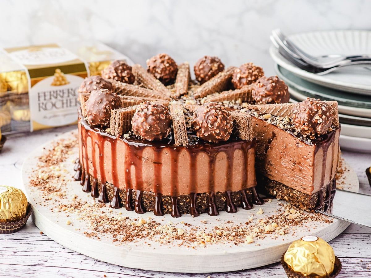 Slicing cheesecake, with Ferrero Rocher and stack of plates and forks in background.