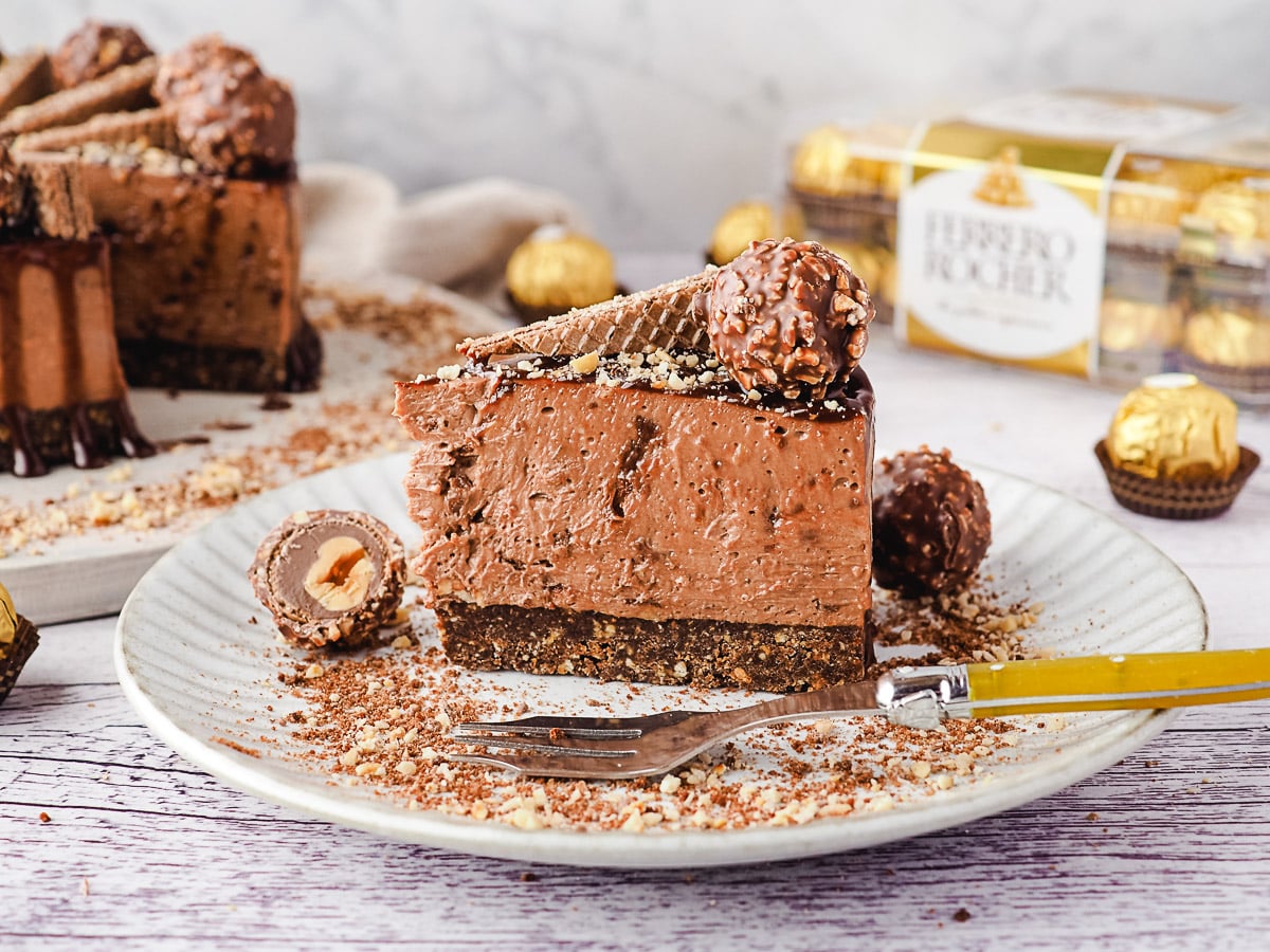 slice of cheesecake on a plate with a fork, with Ferrero Rocher and rest of cheesecake in the background.