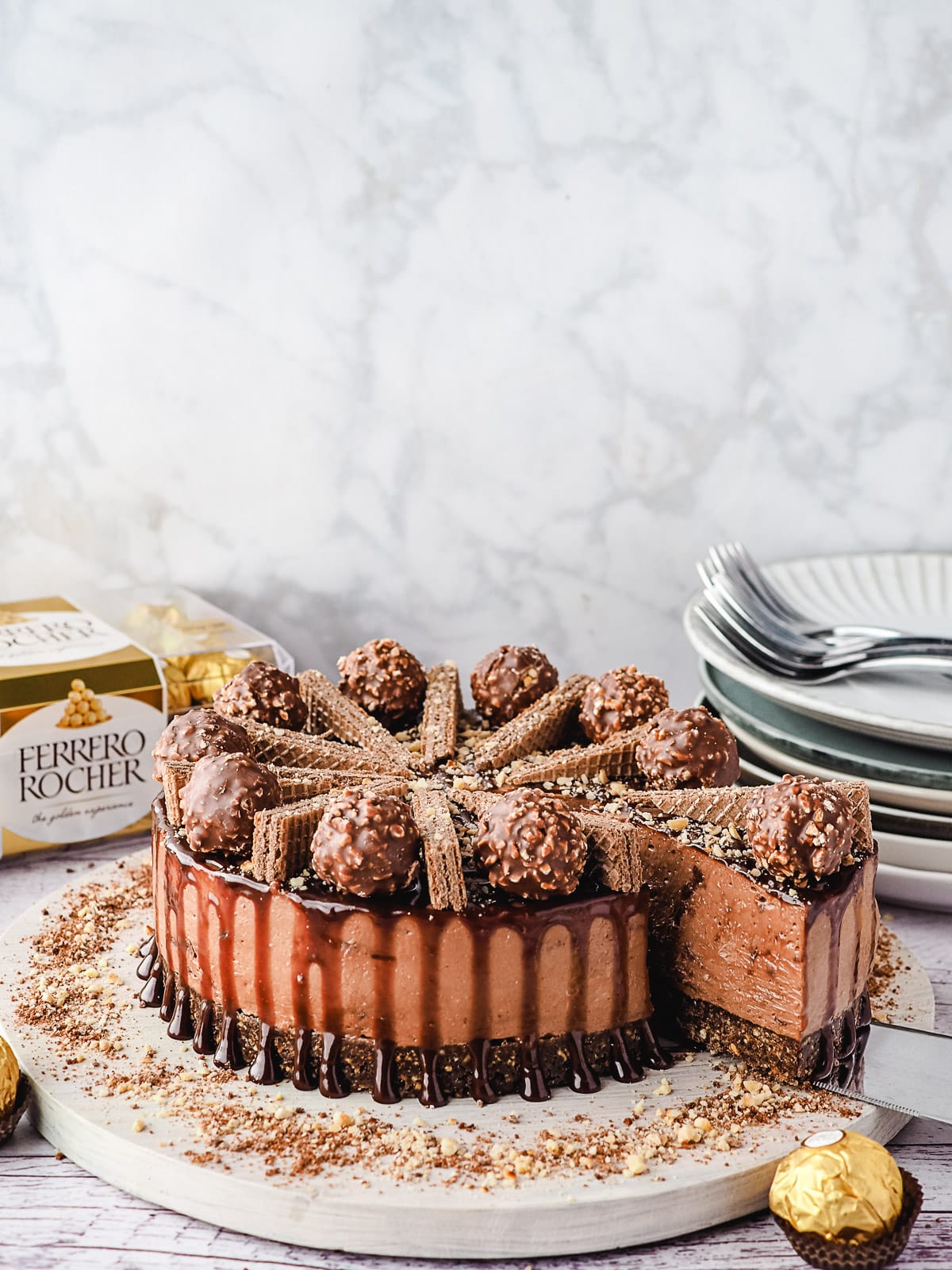 Slicing cheesecake, with Ferrero Rocher and stack of plates and forks in background.