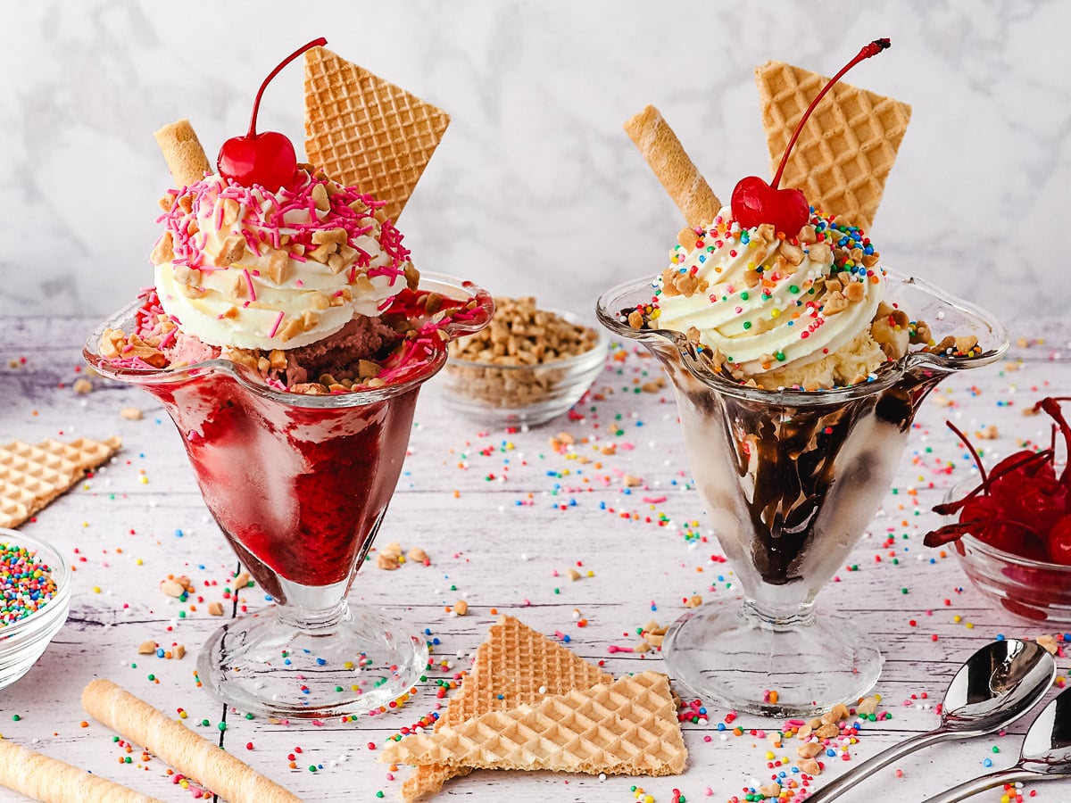 Strawberry and classic sundaes in sundae cups, surrounded by sprinkles, wafers, spoons, nuts and maraschino cherries.