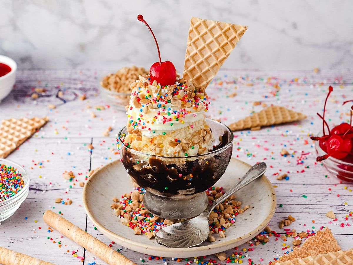 Classic sundaes in a cup, surrounded by sprinkles, wafers, spoons, nuts and maraschino cherries.