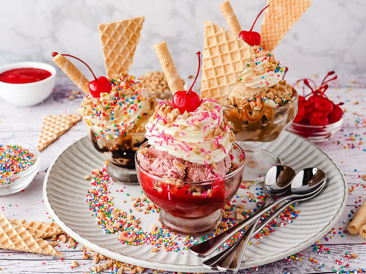 Group of three sundaes, strawberry, classic and caramel, with whipped cream, nuts, sprinkles, wafers and cherry, on a plate with spoons, nuts and sprinkles.