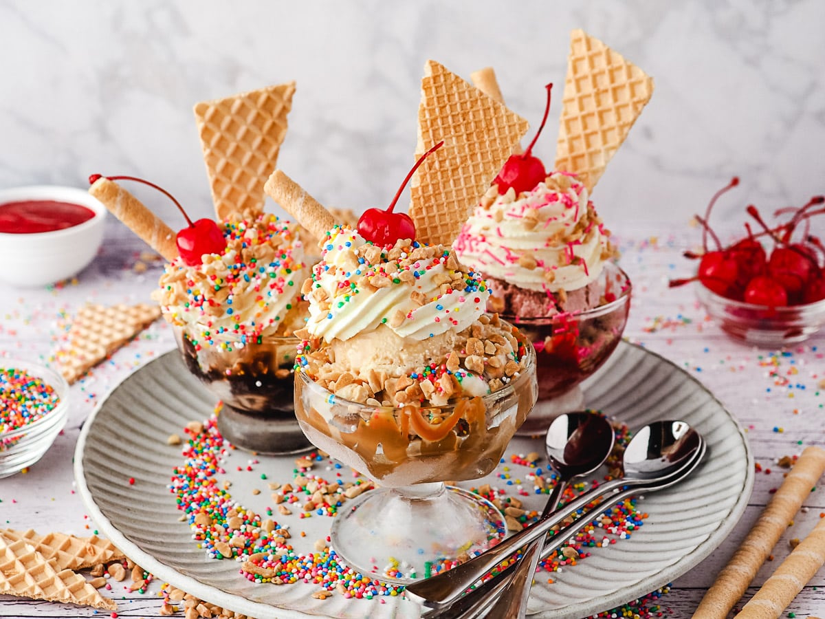 Group of three sundaes, caramel, classic and strawberry, with whipped cream, nuts, sprinkles, wafers and cherry, on a plate with spoons, nuts and sprinkles.