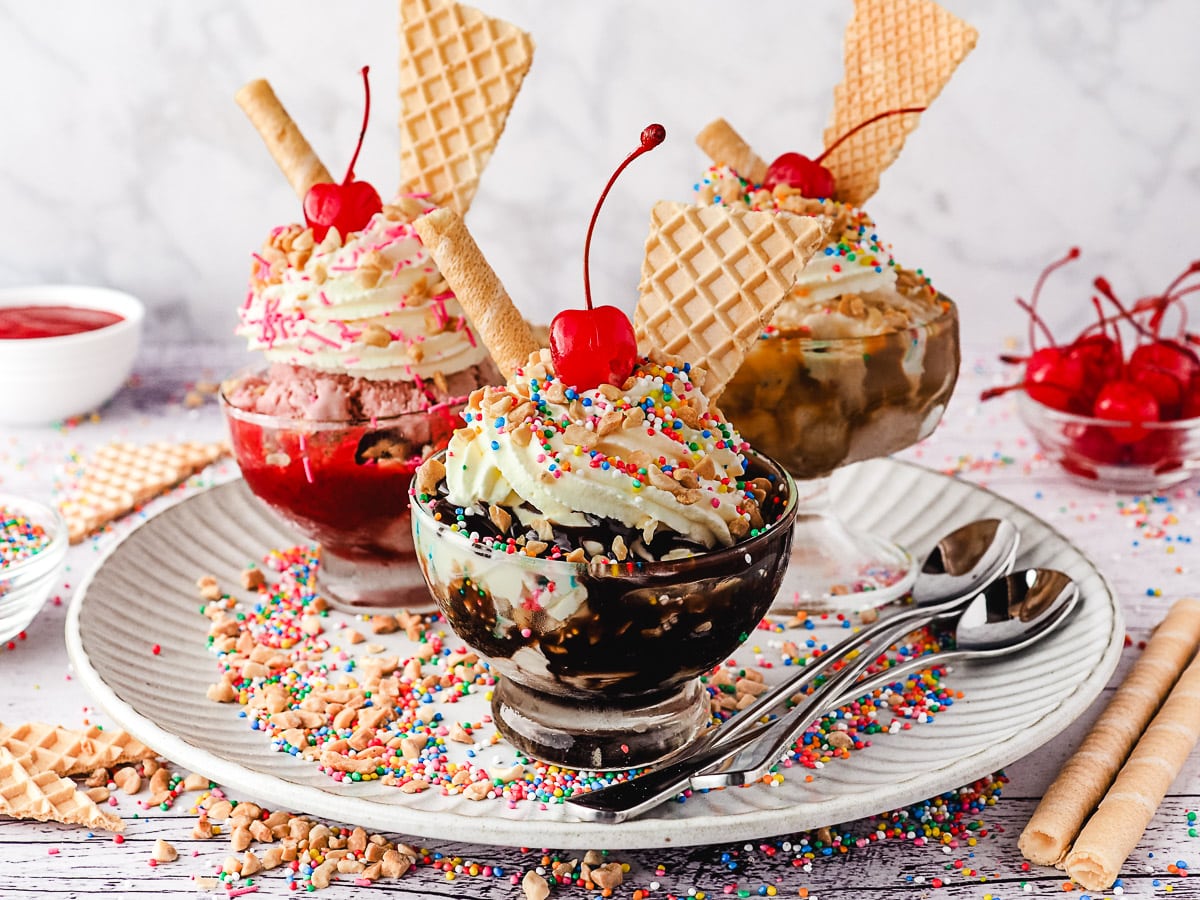 Group of three sundaes, classic, strawberry and caramel, with whipped cream, nuts, sprinkles, wafers and cherry, on a plate with spoons, nuts and sprinkles.