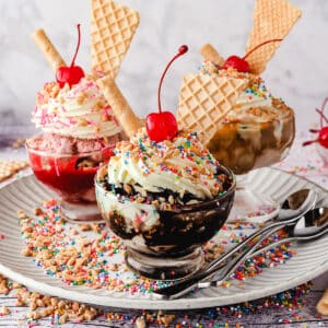 Group of three sundaes, classic, strawberry and caramel, with whipped cream, nuts, sprinkles, wafers and cherry, on a plate with spoons, nuts and sprinkles.